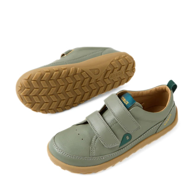 The Bobux KP Kids Plus Dawn Leather Sneaker in Vetiver Green features a barefoot-inspired design with tan rubber zero-drop soles, two Velcro straps, and a teal pull tab at the heel. One sneaker is shown upright while the other displays its tread pattern and wide toe box, both crafted to encourage natural foot movement.