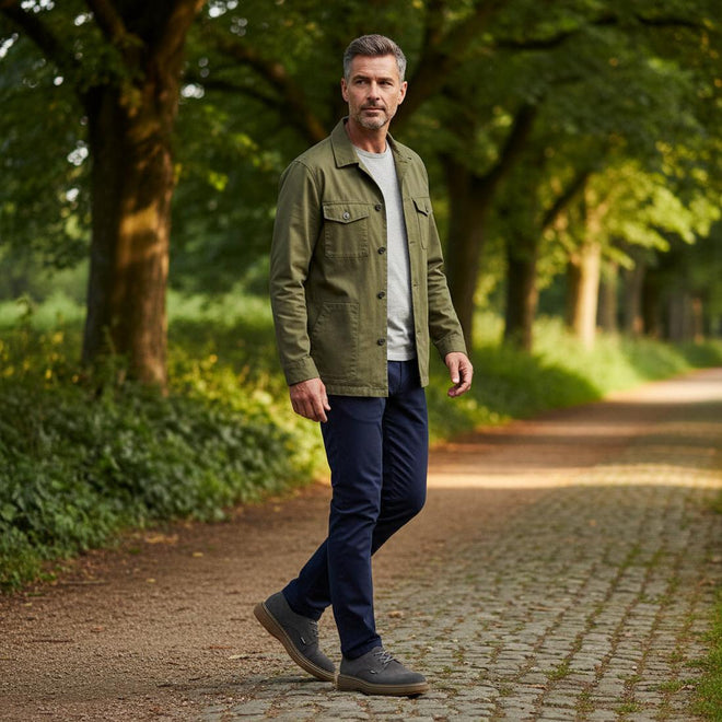 A man in a tree lined path wearing navy trousers a green jacket and grey shirt waering The Barbour Mens Harefield Derby Lace Up Shoe in slate grey suede features brown rubber soles and is displayed on a white background.