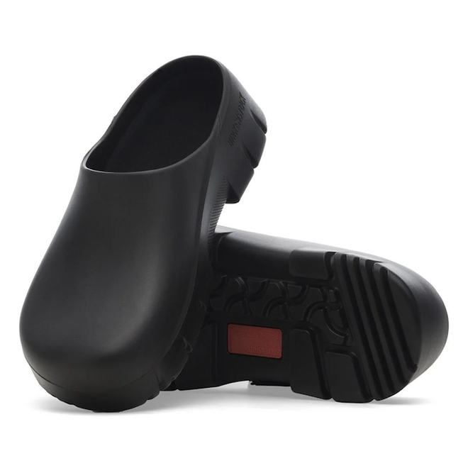 The BIRKENSTOCK Super Birki 2.0 Polyurethane Black clogs feature thick, textured, slip-resistant soles and a red detail—ideal waterproof shoes for the kitchen or garden. One clog stands upright; the other displays the patterned grip.