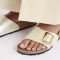 Close-up of a person wearing Birkenstock Womens Catalina Cushion Buckle Birko Flor Pearl White sandals (Regular Width) with a signature cork-latex footbed and large gold buckle, paired with light pants on a white surface., 4066648995265, 4066648995302, 4066648995340, 4066648995388, 4066648995425, 4066648995463, 47502