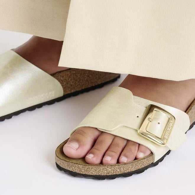 Close-up of a person wearing Birkenstock Womens Catalina Cushion Buckle Birko Flor Pearl White sandals (Regular Width) with a signature cork-latex footbed and large gold buckle, paired with light pants on a white surface., 4066648995265, 4066648995302, 4066648995340, 4066648995388, 4066648995425, 4066648995463, 47502