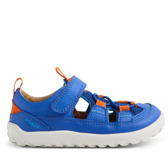 The Bobux IW Iwalk Rocky Dazzling Blue Closed Toe Sandal Barefoot is a kids’ sandal in blue and orange, with a barefoot-shaped toe box, white rugged sole, adjustable Velcro strap, and breathable cutout sides, shown from the side.