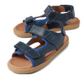 The Bobux Kid+ Plus Strand Navy Open Toe Sandal features adjustable Velcro straps, tan rubber soles, and a breathable design, providing comfort and casual style for kids.
