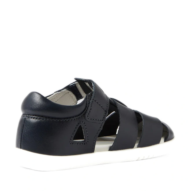 A navy Bobux IWalk Tidal Closed Toe Quick Dry Sandal (634404A), featuring cut-out sides, a closed heel, and a white sole, is shown from the back left angle against a white background.