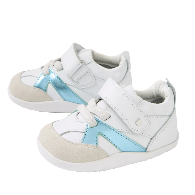 A pair of first walking toddler sneakers in white, light beige suede, and metallic light blue, featuring hook-and-loop straps and elastic laces, shown on a white background.