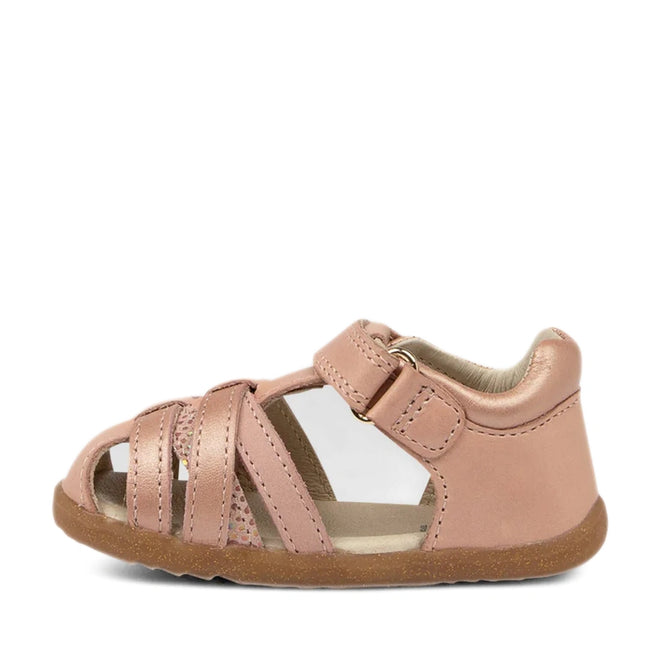 The Bobux Step Ups Cross Jump Dusk Pearl Pink Closed Toe Sandal is a pink toddler sandal featuring a closed heel, open toe, and multiple straps across the front. It has a Velcro ankle strap and a brown rubber sole., 9420062083222, 9420062083239, 9420062083246, 9420062083253