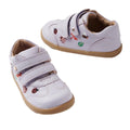 The Bobux Iwalk Buggie Thistle Lavender Embroidered Trainer is a pair of white toddler shoes with tan soles, colorful animal and nature designs, and two velcro straps on each shoe.