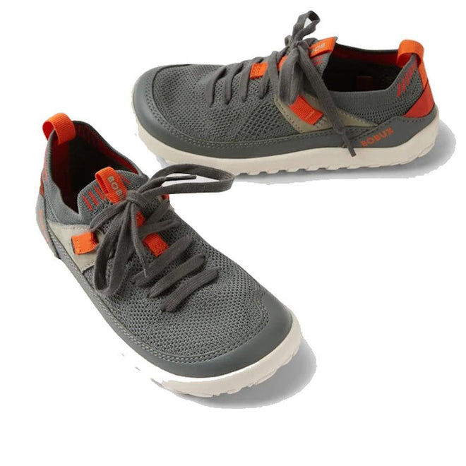 The Bobux Kid+ Meta Knit Barefoot Sneaker in Vetiver Green for juniors features orange accents, white soles, a breathable knit upper, and gray laces, shown on a white background.