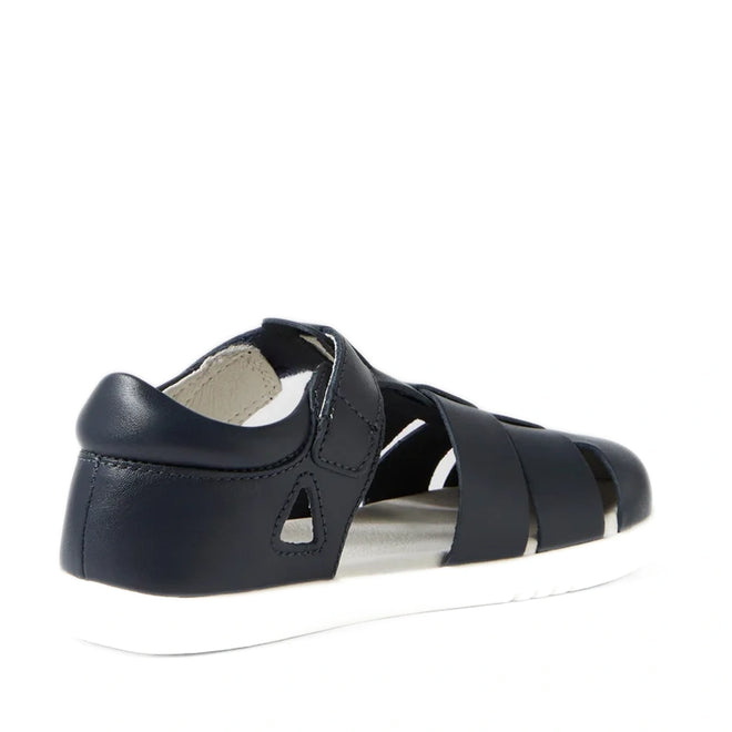 The Bobux Kid+ Plus Tidal Closed Toe Quick Dry Sandal Navy 834404A is shown from the back, featuring a navy blue design with a closed heel, open sides, and a contrasting white sole. This sandal has a closed toe, secure Velcro strap, and multiple horizontal bands across the upper for enhanced comfort and support.