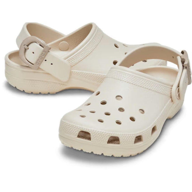 The Crocs Adults Classic Enamel Buckle Clog Frappe features a beige slip-on design with ventilation holes, adjustable back straps, and signature Croslite foam for comfort, displayed against a white background.
