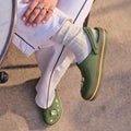 A person in cream pants with a black stripe, cream socks, and Crocs Adults Crocband Gum Clog Cargo Green with Croslite foam charms sits by a round table on sunlit pavement, their ring-adorned hand resting on their knee., 51556, 198445373183, 198445373190, 198445373206, 198445373213, 198445373220, 198445373138, 198445373145, 198445373152, 198445373169