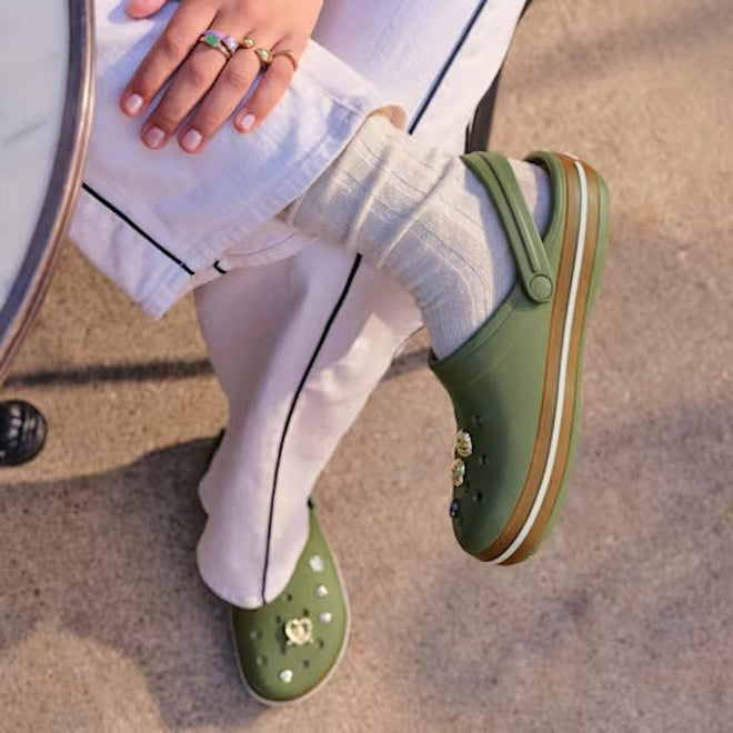 A person in cream pants with a black stripe, cream socks, and Crocs Adults Crocband Gum Clog Cargo Green with Croslite foam charms sits by a round table on sunlit pavement, their ring-adorned hand resting on their knee., 51556, 198445373183, 198445373190, 198445373206, 198445373213, 198445373220, 198445373138, 198445373145, 198445373152, 198445373169