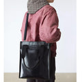 Persond back view holding a large Dr Martens Black tote bag over the shoulder , showing the metal logo on the front,  shoulder strap and handles with a plain background.,
190665739572