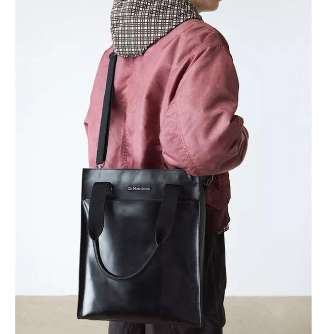 Persond back view holding a large Dr Martens Black tote bag over the shoulder , showing the metal logo on the front,  shoulder strap and handles with a plain background.,
190665739572