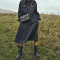 A person stands on grassy terrain under cloudy skies, wearing a dark denim shirt and midi skirt, and carrying a small crossbody bag. They complete the look with Barbour Devlin Black Mid Calf Wellys Womens Rainboot featuring floral designs., 51446, 5063669171547, 5063669171554, 5063669171561, 51450, 51451