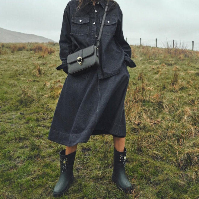 A person stands on grassy terrain under cloudy skies, wearing a dark denim shirt and midi skirt, and carrying a small crossbody bag. They complete the look with Barbour Devlin Black Mid Calf Wellys Womens Rainboot featuring floral designs., 51446, 5063669171547, 5063669171554, 5063669171561, 51450, 51451