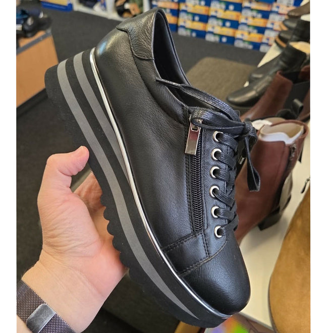 A hand holds the Django & Juliette Eammes Black Leather Platform Shoe Women's, featuring a thick striped sole, side zipper, and laces. Shoe shelves with various footwear boxes are visible in the background., 51089, 4434754993411, 4434756993428, 4434754993435, 4434754993442, 4434754993459, 51095