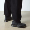 A person in black pants and Dr. Martens 101 UB Black Ambassador 6-Eye Leather Boots with yellow stitching stands on a light floor, highlighting the soft leather footwear and lower legs., 190665693683, 190665693690, 190665693706, 190665693720, 190665693737, 190665693744, 190665693768, 190665693775