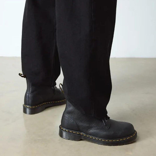 A person in black pants and Dr. Martens 101 UB Black Ambassador 6-Eye Leather Boots with yellow stitching stands on a light floor, highlighting the soft leather footwear and lower legs., 190665693683, 190665693690, 190665693706, 190665693720, 190665693737, 190665693744, 190665693768, 190665693775