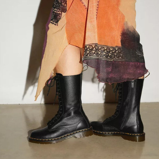 A person wearing a colorful, asymmetrical skirt and black Dr Martens - 1B99 14 Up Zip Black Boot Virginia Leather stands on a beige floor against a white wall.