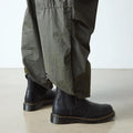 Person in loose olive green pants and Dr. Martens 2976 Ambassador Black Leather Chelsea Boots with Goodyear-welted yellow stitching, standing on a smooth, light-colored floor., 50056, 50057, 50059, 190665663303, 190665663310, 190665663327, 50067, 50068