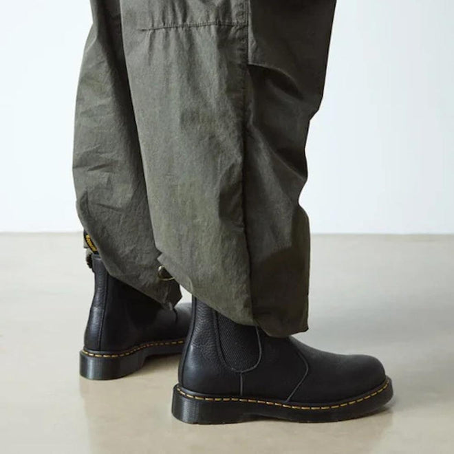 Person in loose olive green pants and Dr. Martens 2976 Ambassador Black Leather Chelsea Boots with Goodyear-welted yellow stitching, standing on a smooth, light-colored floor., 50056, 50057, 50059, 190665663303, 190665663310, 190665663327, 50067, 50068