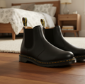 A pair of Dr Martens 2976 Ambassador Black Leather Chelsea Boots with yellow stitching and branded pull tabs sits on a wooden floor in a cozy bedroom with a bed and nightstand in the background., 50056, 50057, 50059, 190665663303, 190665663310, 190665663327, 50067, 50068
