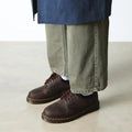 A person in wide-leg olive green pants and Dr Martens 8053 Crazy Horse Dark Brown Leather 5 Eye shoes with yellow stitching stands on a light floor. The unisex shoe has an air-cushioned sole, and the blue coat's lower part is visible., 52072, 52073, 800090801651, 800090801675, 800090801682, 800090801699, 800090801712