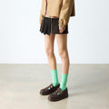 A person stands indoors on a polished concrete floor wearing a tan jacket, black pleated skirt, bright green socks, and dark brown Dr Martens 8065 2 Strap Mary Jane Shoe Crazy Horse Brown. Their face is not visible., 190665588941, 190665588958, 190665588965, 190665588972, 13164654654, 190665588996, 190665589009, 190665589016