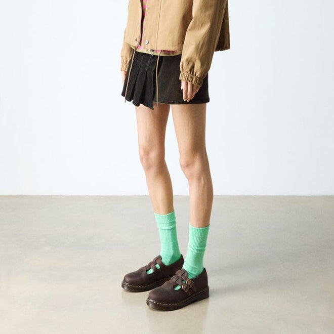 A person stands indoors on a polished concrete floor wearing a tan jacket, black pleated skirt, bright green socks, and dark brown Dr Martens 8065 2 Strap Mary Jane Shoe Crazy Horse Brown. Their face is not visible., 190665588941, 190665588958, 190665588965, 190665588972, 13164654654, 190665588996, 190665589009, 190665589016