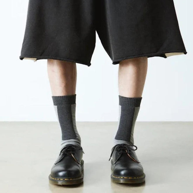 Person wearing black shorts, Dr Martens Socks Double Doc Sock Unisex Charcoal Organic, and black lace-up shoes stands on a light-colored floor against a plain white background., 190665563108, 190665563092