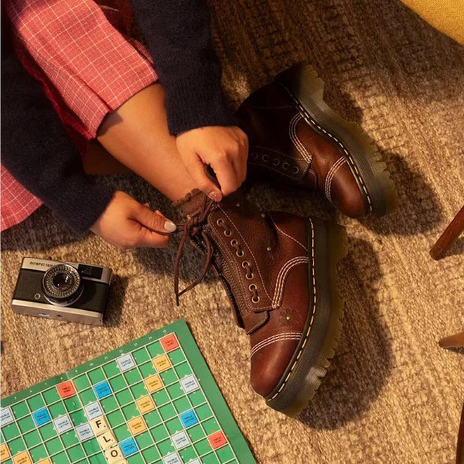Someone wearing pink plaid pants and Dr Martens Sinclair Flower Dark Brown Grizzly Leather Boots with a platform Quad sole ties their laces beside a vintage camera and Scrabble board on a carpeted floor., 51903, 51904, 190665777116, 190665777123, 190665777147, 51909, 51910