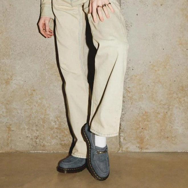 A person in cream pants and white socks wears Dr. Martens Adrian Snaffle Loafers in Washed Denim Tumbled Nubuck, standing against a textured beige wall. Only the lower half of their body is visible.