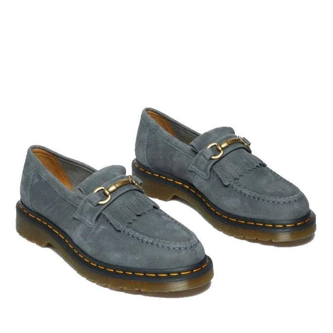 A pair of Dr. Martens Adrian Snaffle Loafers in washed denim tumbled nubuck, featuring fringe detailing, gold horsebit hardware, and signature yellow stitching on thick translucent rubber soles.