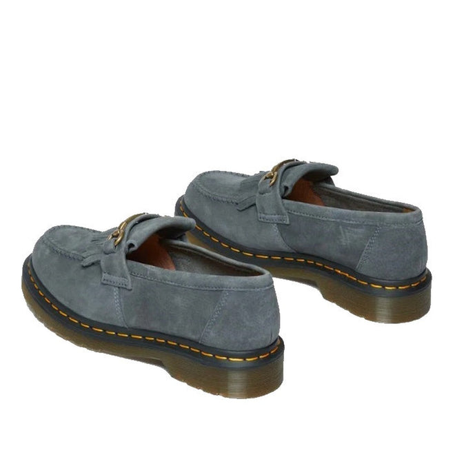 A pair of Dr. Martens Adrian Snaffle Loafers in washed denim tumbled nubuck with gold-tone buckles, yellow stitching, and brown translucent outsoles, shown from a rear side angle on a white background.