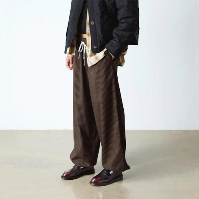 A person stands indoors with hands in pockets, wearing loose brown pinstriped pants, a beige shirt, a black jacket, and Dr Martens Temara Cherry Red Arcadia Leather Loafers. Their face is not visible., 190665747560, 190665747577, 190665747591, 52060, 52061