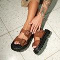 A person with a tattooed arm wears Dr Martens Zebzag Sepia Brown Nubuck 2 Strap Sandals with black soles and gold buckles, sitting on a speckled tile floor. Their ring-and-bracelet adorned hand rests on their foot., 190665790603, 190665790610, 190665790627, 190665790641, 190665790658, 190665790665