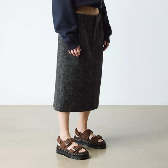 Indoors, a person wears a dark gray knee-length skirt, navy long-sleeve top with extended sleeves, and Dr Martens Zebzag Sepia Brown Nubuck 2 Strap Sandals. The minimal background features a light floor and wall., 190665790603, 190665790610, 190665790627, 190665790641, 190665790658, 190665790665