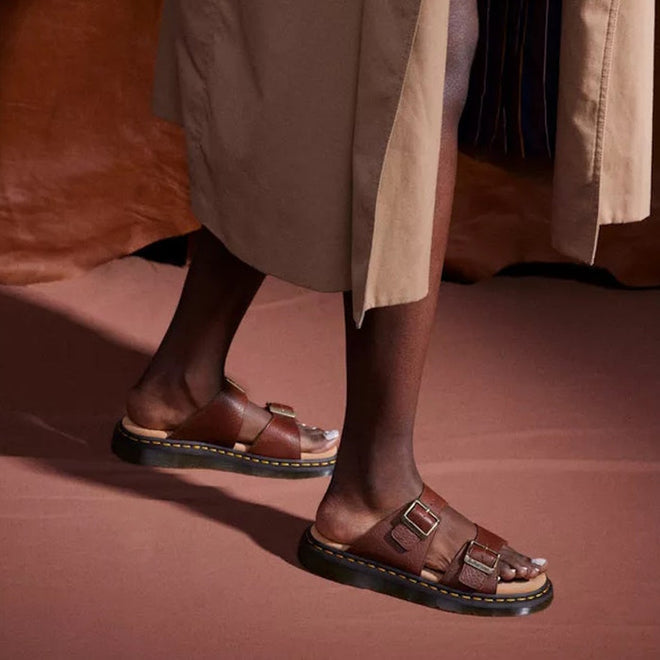 A person wearing a beige trench coat and Dr Martens Josef Slide Sandal in Cashew Ambassador Leather stands on a brown surface, with only their lower legs and feet visible.