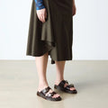 A person wears a dark olive asymmetrical midi skirt and black Dr Martens Josef Charro Atlas Slide Sandal Leather on a light floor, with only the lower half of their body visible.