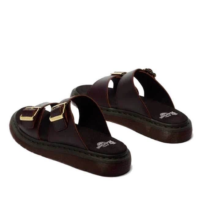 The Dr Martens Josef Charro Atlas Slide Sandal features dark brown Atlas leather, two wide straps with gold buckles, an open toe design, and a thick textured sole, shown against a white background.