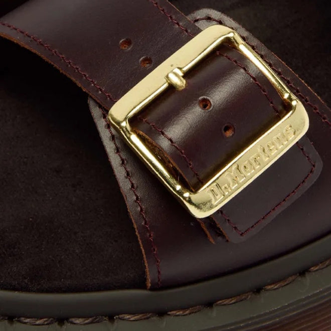 Close-up of the Dr Martens Josef Charro Atlas Slide Sandal Leather, featuring a brown leather strap with a gold buckle engraved with Dr. Martens, detailed stitching, and part of the black Goodyear-welted sole.