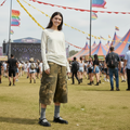 A woman in a white long-sleeve shirt, camo cargo shorts, striped socks, and Dr Martens Madaline Black Smooth Womens Slingback Mary Jane Shoes smiles at an outdoor music festival amid colorful tents and a lively crowd.