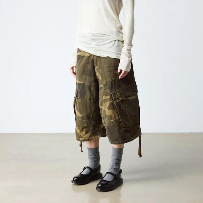 A person wearing a long-sleeve white top, loose camouflage cargo shorts, gray socks, and Dr. Martens Madaline Black Smooth Womens Slingback Mary Jane Shoes stands indoors against a plain light background. The person's head is not visible.