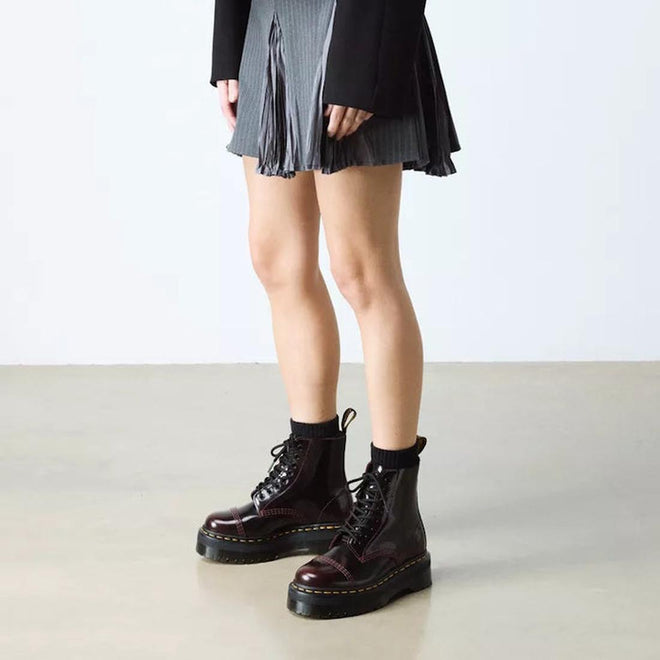 A person wearing a gray pleated skirt, black blazer, black socks, and Dr Martens Sinclair Cherry Red Arcadia Leather Platform Boots stands on a smooth beige floor against a plain white background., 51252, 190665278637, 190665278644, 190665278668, 51256
