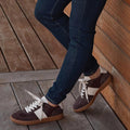 Wearing dark jeans and EOS Antigo Cacao Brown Leather Lace Up Trainers, featuring white stripes and gum soles, standing on a wooden floor and leaning against a wooden wall. Only legs and shoes are visible., 51643, 9321191255156, 9321191255163, 9321191255170, 9321191255187, 9321191255194, 51649