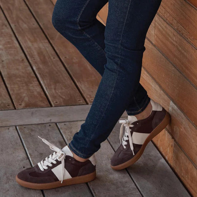 Wearing dark jeans and EOS Antigo Cacao Brown Leather Lace Up Trainers, featuring white stripes and gum soles, standing on a wooden floor and leaning against a wooden wall. Only legs and shoes are visible., 51643, 9321191255156, 9321191255163, 9321191255170, 9321191255187, 9321191255194, 51649