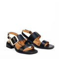 The EOS Ikam Women's Black Leather Low Heeled Sandal features wide crisscross straps, a back strap, tan insoles, large gold square buckles, and a low block heel, shown against a plain white background., 9321191304915, 9321191304922, 9321191304939, 9321191304946, 9321191304953