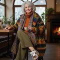 Woman sitting in a cozy room with a fireplace, wearing a colorful coat and holding a cup. Wearing EOS Jovi sneakers in Pony haire with white, brown, black camo like bovine pattern.,  9321190036015, 9321190036022, 9321190036039, 9321190036046, 9321190036053, 51488, 51482, 9321190036015, 9321190036022, 9321190036039, 9321190036046, 9321190036053, 51488