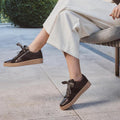A person sits outdoors on a chair, one foot slightly raised, wearing wide-legged white pants and EOS Tinah Mahogany Dark Brown Leather Trainers With Zip., 51468, 9321190042108, 9321190042115, 9321190042122, 9321190042139, 9321190042146, 51474